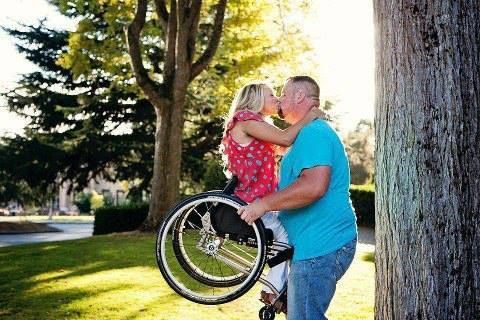 Un hombre levanta a su mujer que va en silla de ruedas para darle un beso