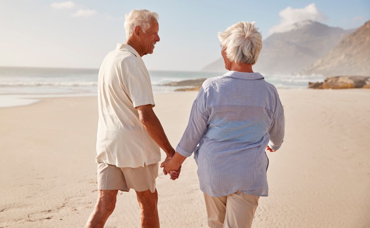 Pareja de personas mayores pasean por una playa durante un viaje del IMSERSO